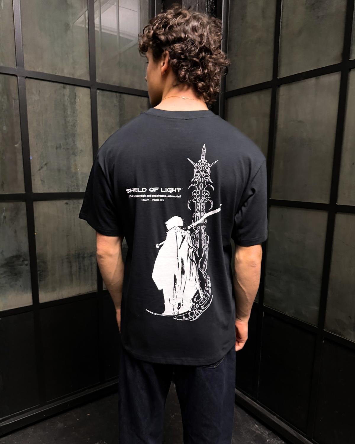 Shield Of Light faith based t-shirt black back design worn by model indoor setting